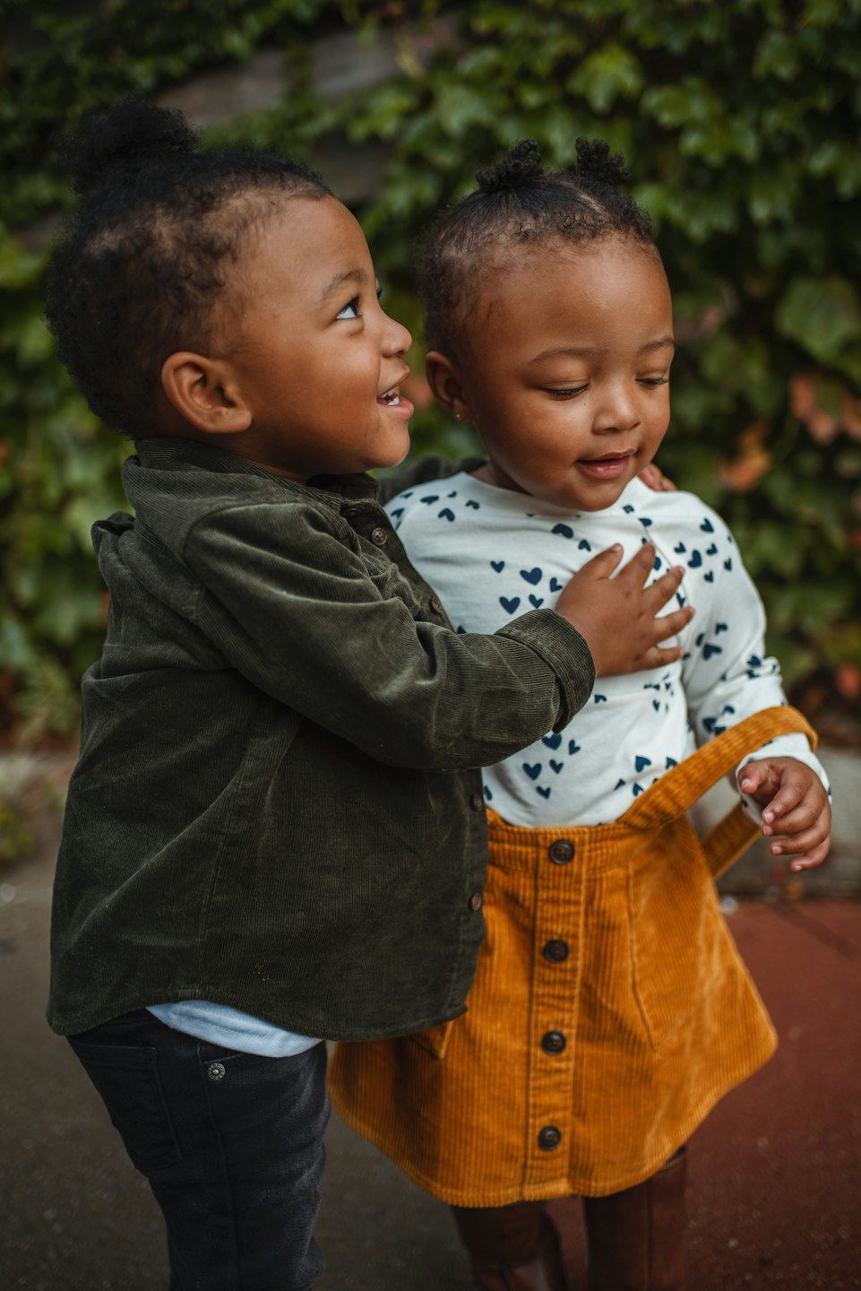 Two smiling children hugging outside near an ivy-covered wall.