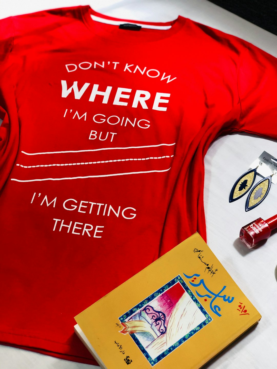 Flat lay of a red t-shirt, book, earrings, and nail polish.